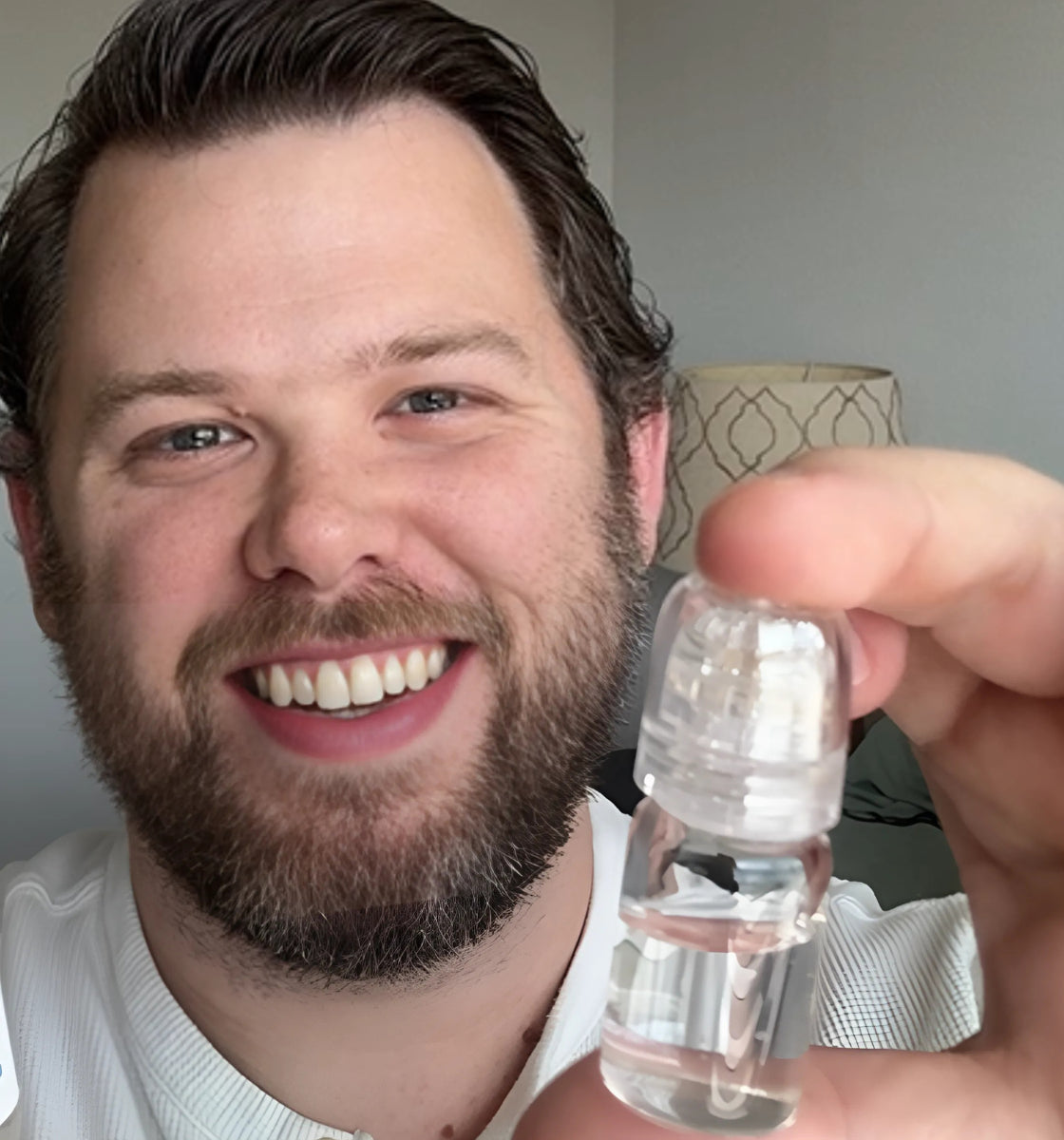 A person smiling while holding a small bottle of clear liquid.